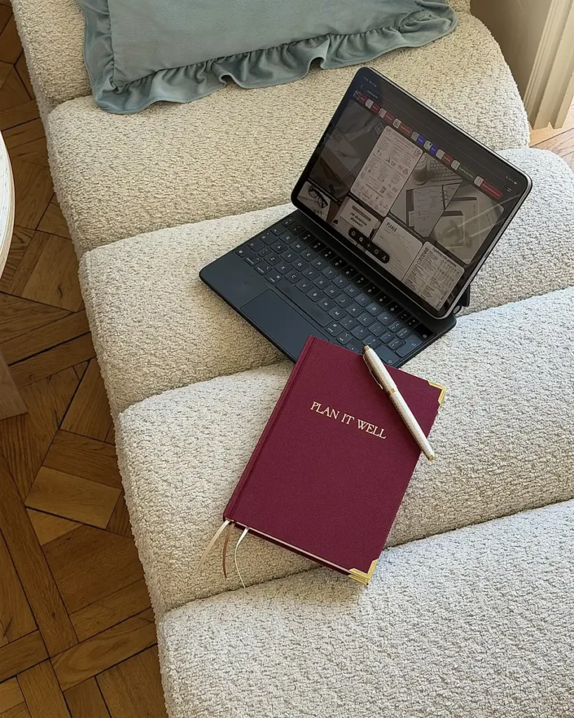 Bordowy planer Plan It Well z długopisem na fotelu z tkaniny boucle, obok tablet